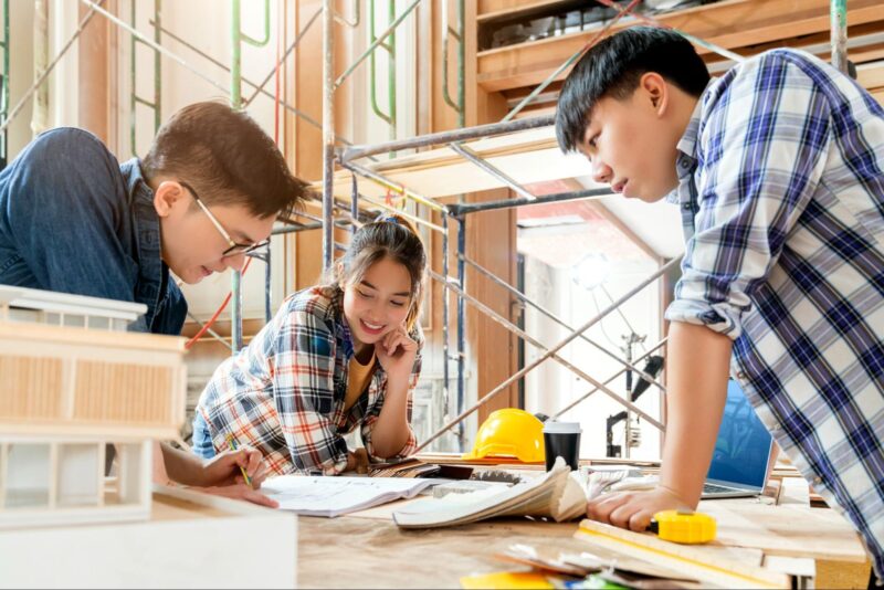 Three architects review building plans and a scale model together at a construction site