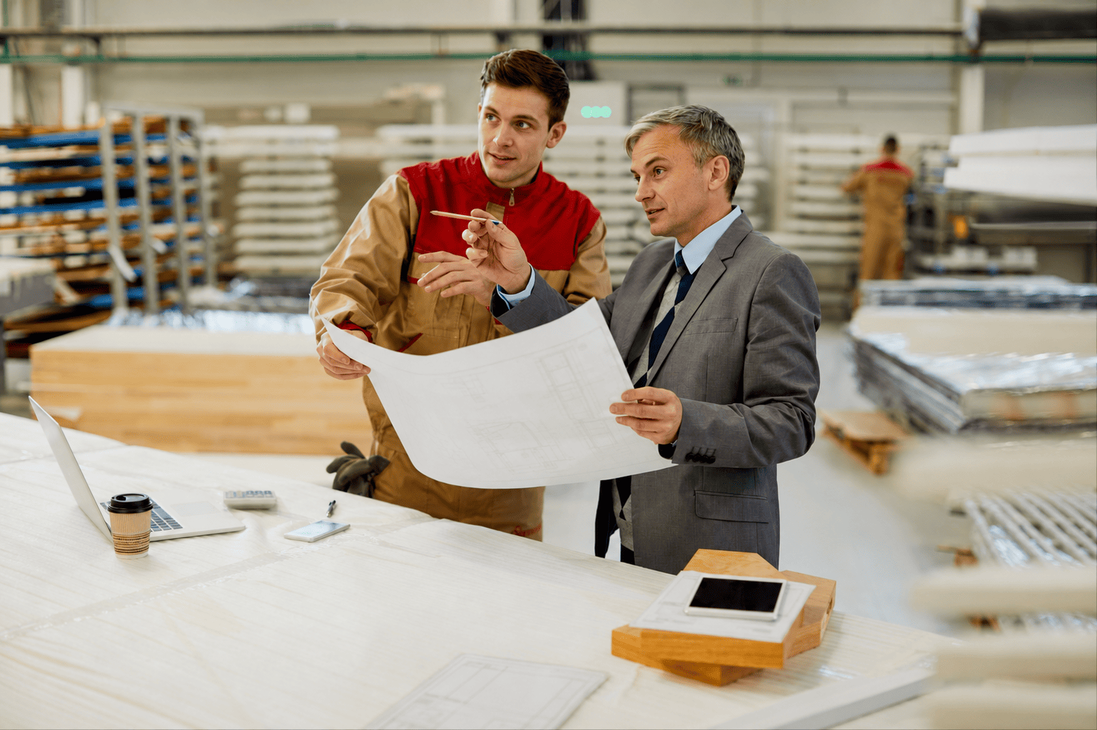 A manager and a worker in a workshop reviewing construction plans together, surrounded by wooden materials and equipment