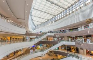 Interior view of a multi-level luxury shopping mall with a skylight ceiling, escalators, and decorative hanging sculptures above retail stores.