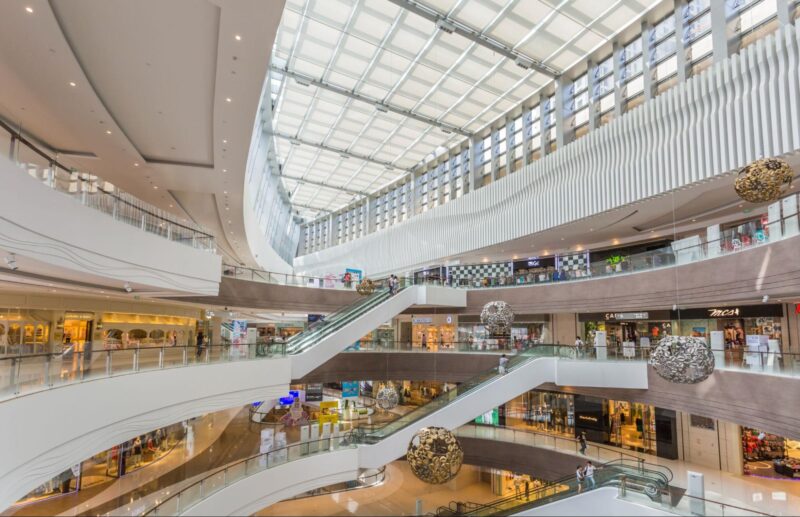 Interior view of a multi-level luxury shopping mall with a skylight ceiling, escalators, and decorative hanging sculptures above retail stores.