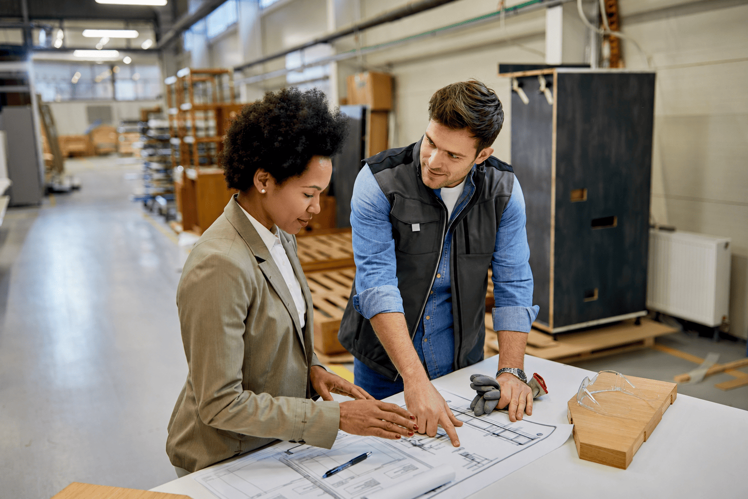 A businesswoman and a warehouse worker review blueprints together on a table at an industrial facility